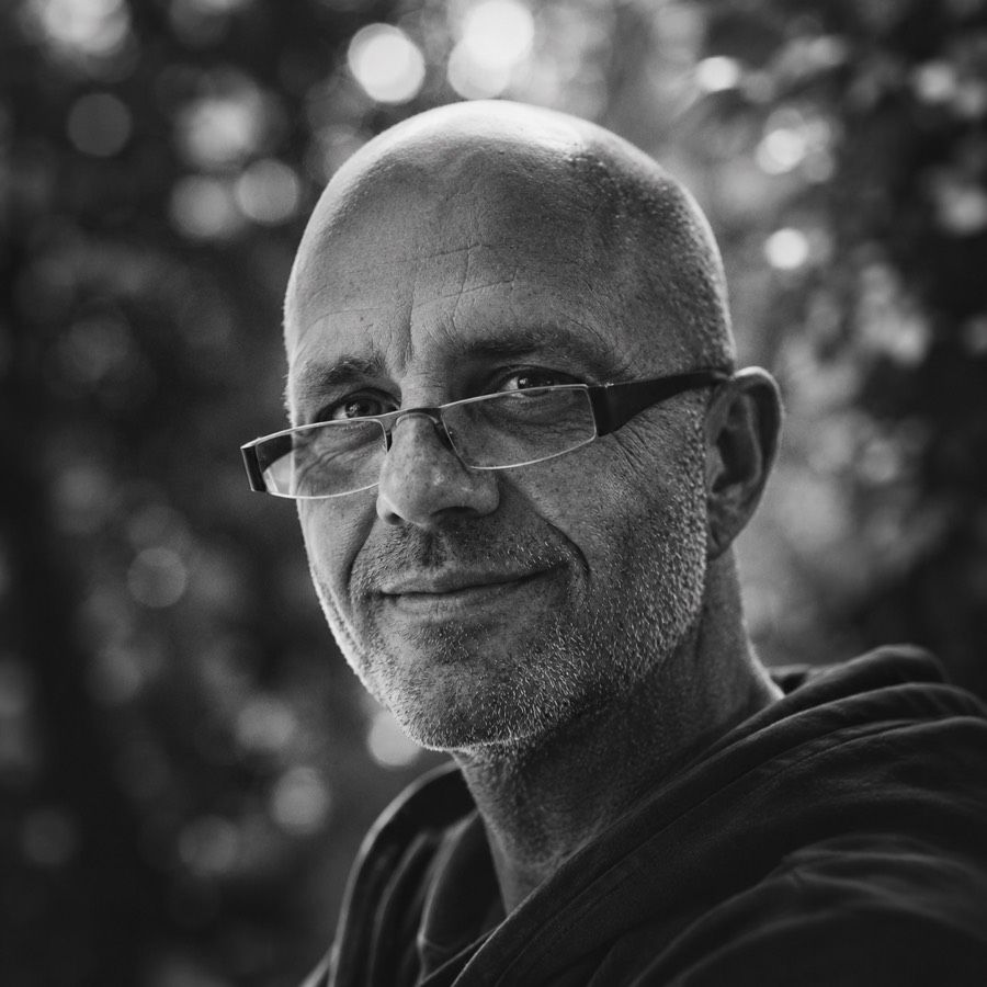 Oliver Wattenberg im Halbprofil, mit Brille und der Hand am Kinn, nachdenklicher Blick, Schwarz-Weiß-Aufnahme im Gegenlicht.