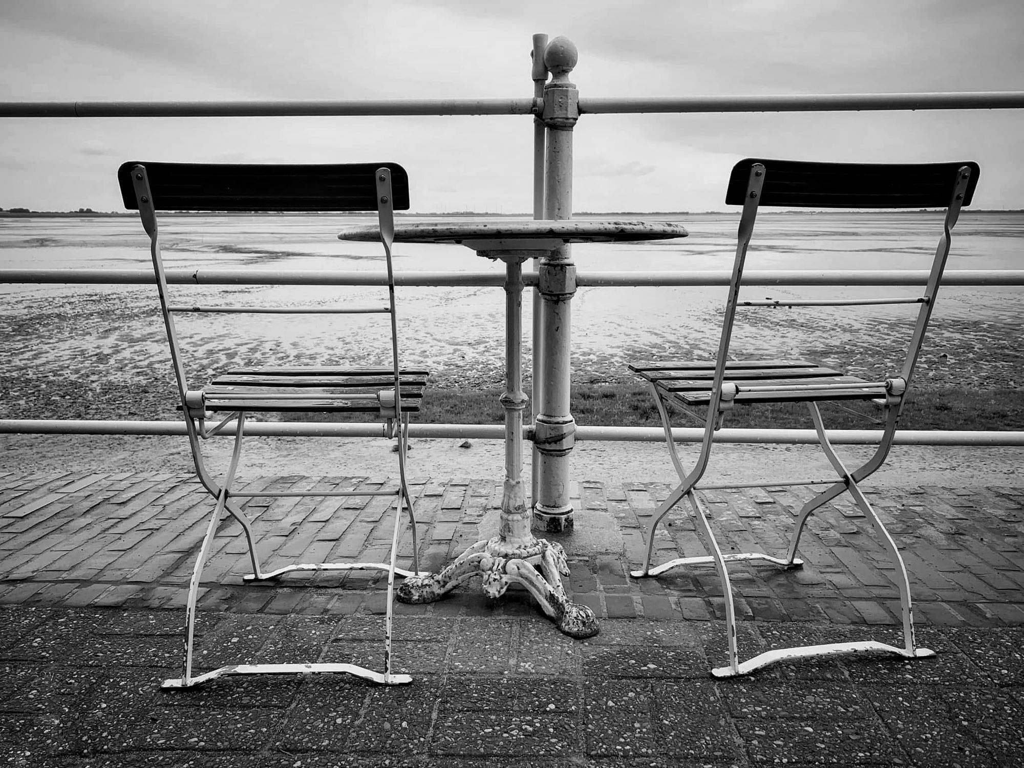 Zwei leere Stühle an einem Tisch an der Promenade, im Hintergrund das Watt bei Ebbe.