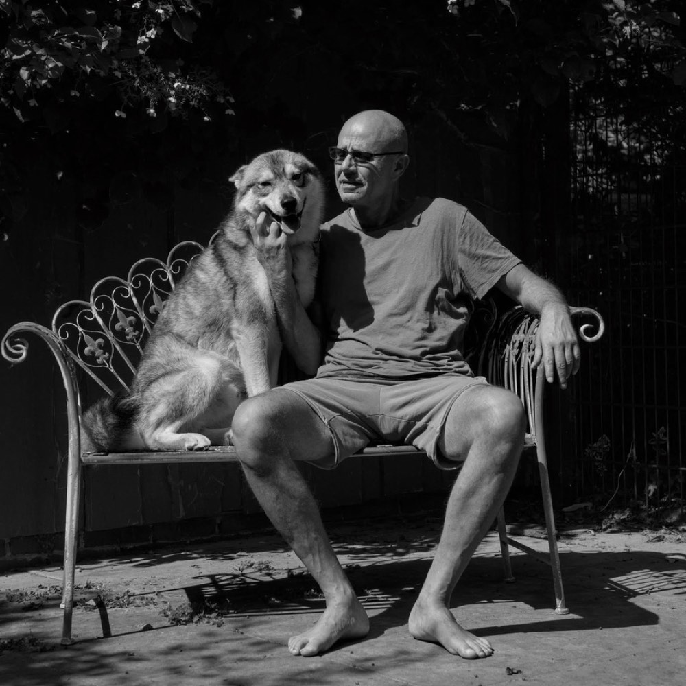 Oliver Wattenberg sitzt barfuß auf einer Gartenbank, neben ihm sein Wolfshund.